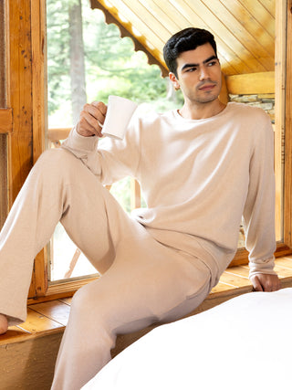 A man wearing a beige cashmere loungewear set with a white mug in hand, posing on a wooden windowsill.
