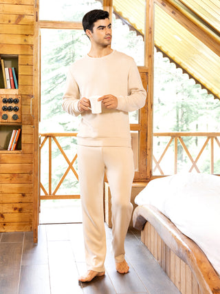 A man in cream-colored loungewear, holding a mug, stands near a wooden cabin window and a bed.