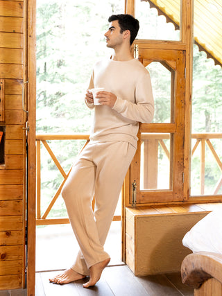 A man in cream-colored loungewear, holding a mug, stands near a wooden cabin window and a bed.
