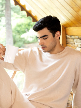 Men's light beige crew neck long sleeve pajamas, posed sitting down with a mug.