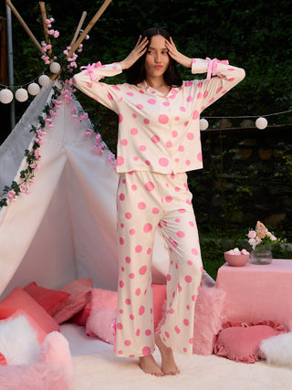 A woman poses playfully in a cream and pink polka dot pajama set with bows on the sleeves and pant legs.