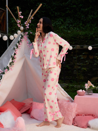 Women in a polka dot pajama set, standing with a hand on hip and finger to chin, near a teepee and pink pillows.