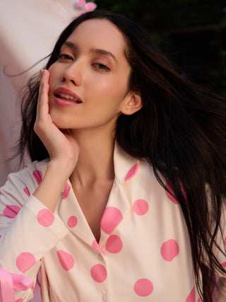 A woman in a cream-colored polka dot pajama set, with pink polka dots and pink ribbon details, gently touches her cheek.