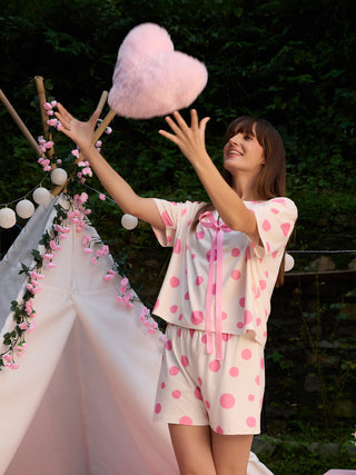 A woman stands in a cream polka dot sleepwear set with pink accents, tossing a pink fluffy heart pillow in the air.