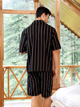 Men's black and white vertical striped short sleeve shirt and shorts loungewear set, viewed from the back, standing by a window with trees outside.