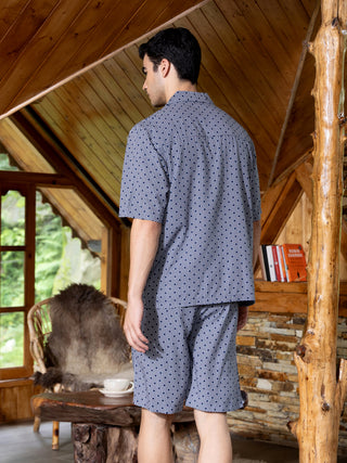 Men's navy blue patterned sleepwear set, model from the back, standing in a rustic wooden cabin with a vintage armchair and a coffee cup on a log table.