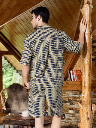 Men's short-sleeved button-down shirt and shorts set in navy blue with a tan geometric print, posed standing with a hand on a wooden pillar.