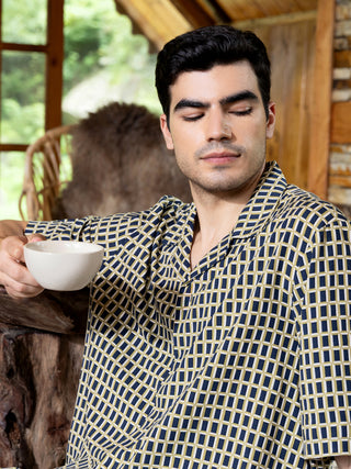 Men's short-sleeved button-down shirt and shorts set in navy blue with a tan geometric print, posed sitting with a coffee cup.