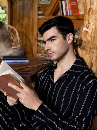 A man in black and white pinstripe sleepwear, sitting and reading a book, with a furry chair in the background.