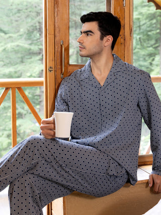 A man in blue printed sleepwear holding a white mug sits by a window.