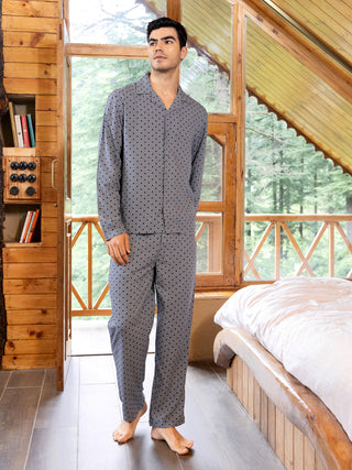 Men posing in a long-sleeved, patterned pajama set with a black and white geometric print, standing indoors by a bed.
