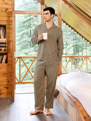 A man in navy blue and yellow printed pajamas holds a white mug while standing near a window.