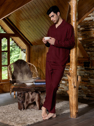 A man in burgundy pajama set with a white polka dot print, holding a cup and leaning against a wooden pillar.