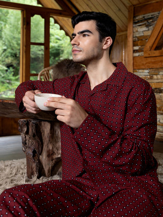 Men wearing a burgundy printed pajama set, holding a cup and looking out a window.
