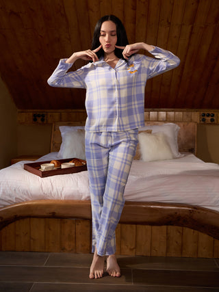 Women wearing a light purple plaid pajama set with a cartoon graphic on the chest, posing with hands on cheeks by a bed.