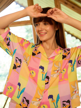 Woman in a pink, yellow, and purple pastel checkered pajama set featuring a Looney Tunes print, striking a playful pose with hands near her head.