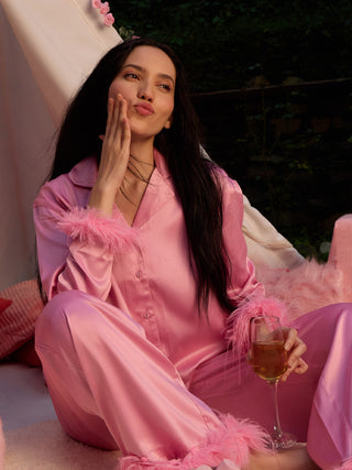 A woman wearing pink silk pajamas with feather trim poses playfully while holding a glass of champagne.