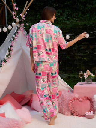 Women's pink pajama set with a playful "Pajama Party" print, featuring various icons and text, shown from the back as she reaches for a string of lights.