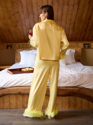 A woman in a yellow silk pajama set with feather trim holds a glass of red wine while standing in a bedroom.