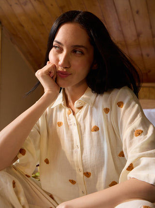 A woman in cream-colored sleepwear with a teddy bear print rests her chin on her hand, looking away from the camera, against a wooden background.