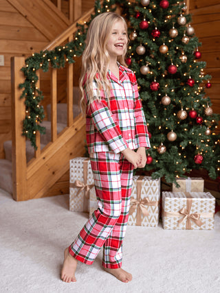 A young girl in a red plaid pajama set poses happily in front of a Christmas tree.