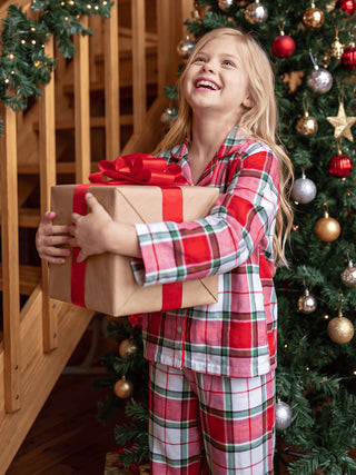 GIRLS wearing red and green plaid pajamas, holding a gift and standing by a Christmas tree.