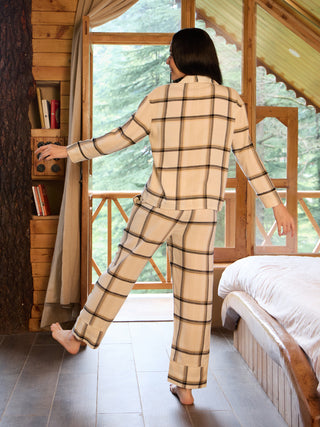 Women in a relaxed pose, wearing cream-colored plaid pajamas, are standing by an open door with a forest view and a bed nearby.