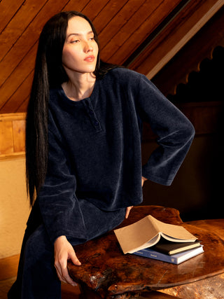 A woman in a navy blue velour pajama set with a relaxed fit poses by a wooden table with books.