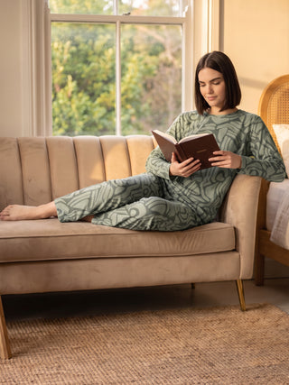 A woman lounges on a tan sofa wearing light green patterned sleepwear, reading a book.