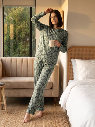 A woman in green abstract print pajamas poses with a mug in her hand, leaning against a wall next to a bed.