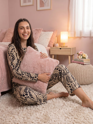 A woman sits on a fluffy rug in a bedroom, holding a pink pillow and wearing a long-sleeved pajama set with a black and beige abstract print.