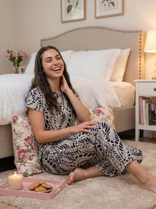 A woman in a navy blue and white patterned sleepwear set sits on the floor with a tray of cookies and a candle.