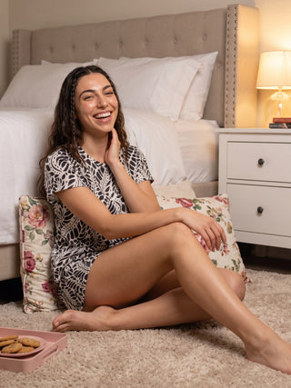 A woman in a black and white printed sleep dress sits on the floor next to a bed.