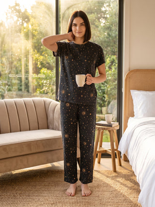A woman stands holding a mug, wearing a dark gray pajama set with a celestial print.