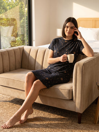 A woman sits on a couch in a dark blue sleep dress with a celestial print, holding a mug.