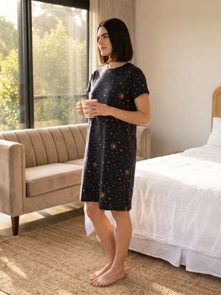 A woman stands in a dark blue sleep dress with a celestial print, holding a mug.