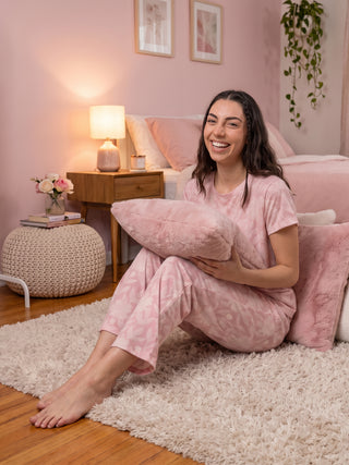 A woman in pink floral print pajamas smiles while holding a pink pillow, sitting on a cream rug.