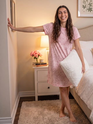 A woman in a pink floral print short-sleeve sleep dress poses with one arm extended to the wall and a white pillow in her other arm.