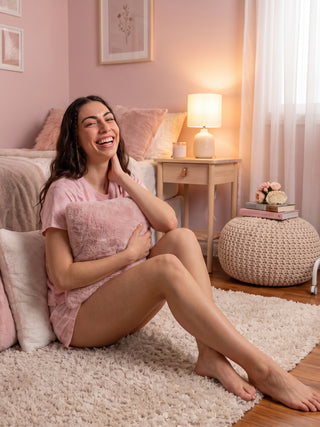 A woman in a pink pajama set is sitting on a rug, holding a pink pillow.