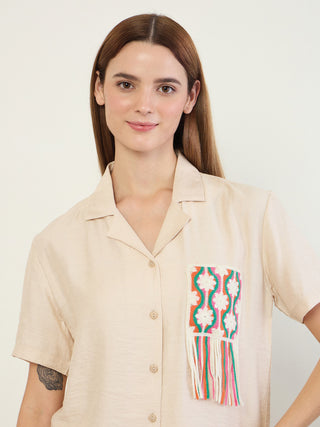A woman poses in a beige pajama top with a colorful embroidered pocket and fringe detail.