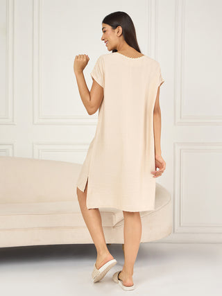 Women in a cream, short-sleeved, midi-length sleep dress with a slit and white slippers, posing with one leg raised.
