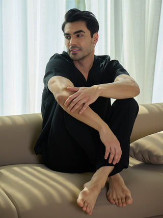 A man in black sleepwear sits cross-legged on a beige sofa, looking away from the camera.