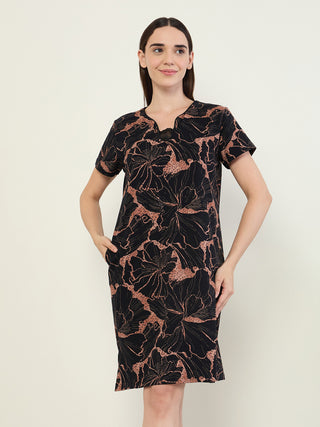 Women in a short-sleeved, black sleep dress with a floral print, standing and looking to the side, with one hand in her pocket.