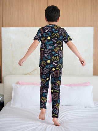 A young boy, seen from behind, energetically bounces on a bed wearing a black sleepwear set featuring a vibrant neon outline print of retro gaming elements and text.
