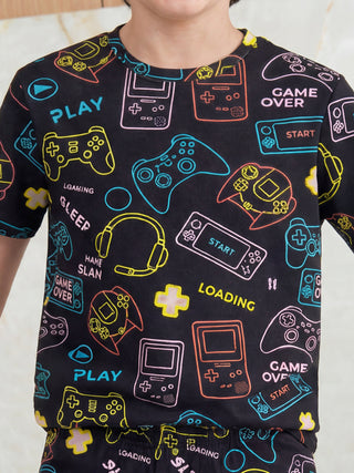 A young person wears a black short-sleeved pajama top adorned with a vibrant neon outline print of gaming controllers, handheld consoles, and playful game-themed text.