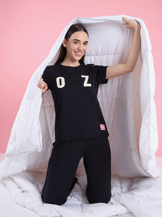 A woman kneels under a white duvet, wearing black pajamas with "OZ" printed in white.