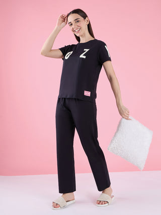 Women's black short-sleeve t-shirt with white "OZ" print and matching black pajama pants, posed standing and holding a fluffy white pillow.