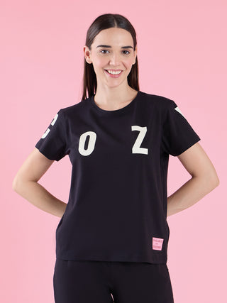 A woman poses in a black graphic t-shirt and matching bottoms, featuring an "OZ" print, with a pink tag on the t-shirt.