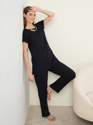 A woman in black pajamas stands leaning against a wall, one leg bent.