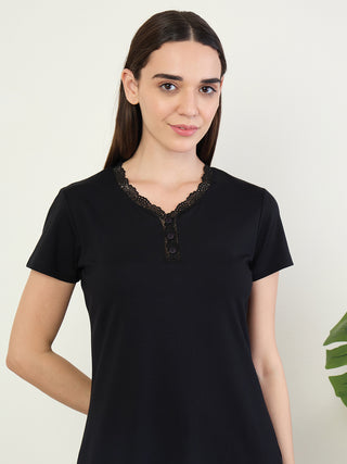 A woman poses for a product shot wearing a black short-sleeved sleep shirt with lace trim and button details.
